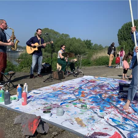 photo d'un atelier de peinture participative, de grand papiers peints par les personnes sont exposés au sol, des musiciens jouent et le public regarde. la scène se passe dans la nature.  