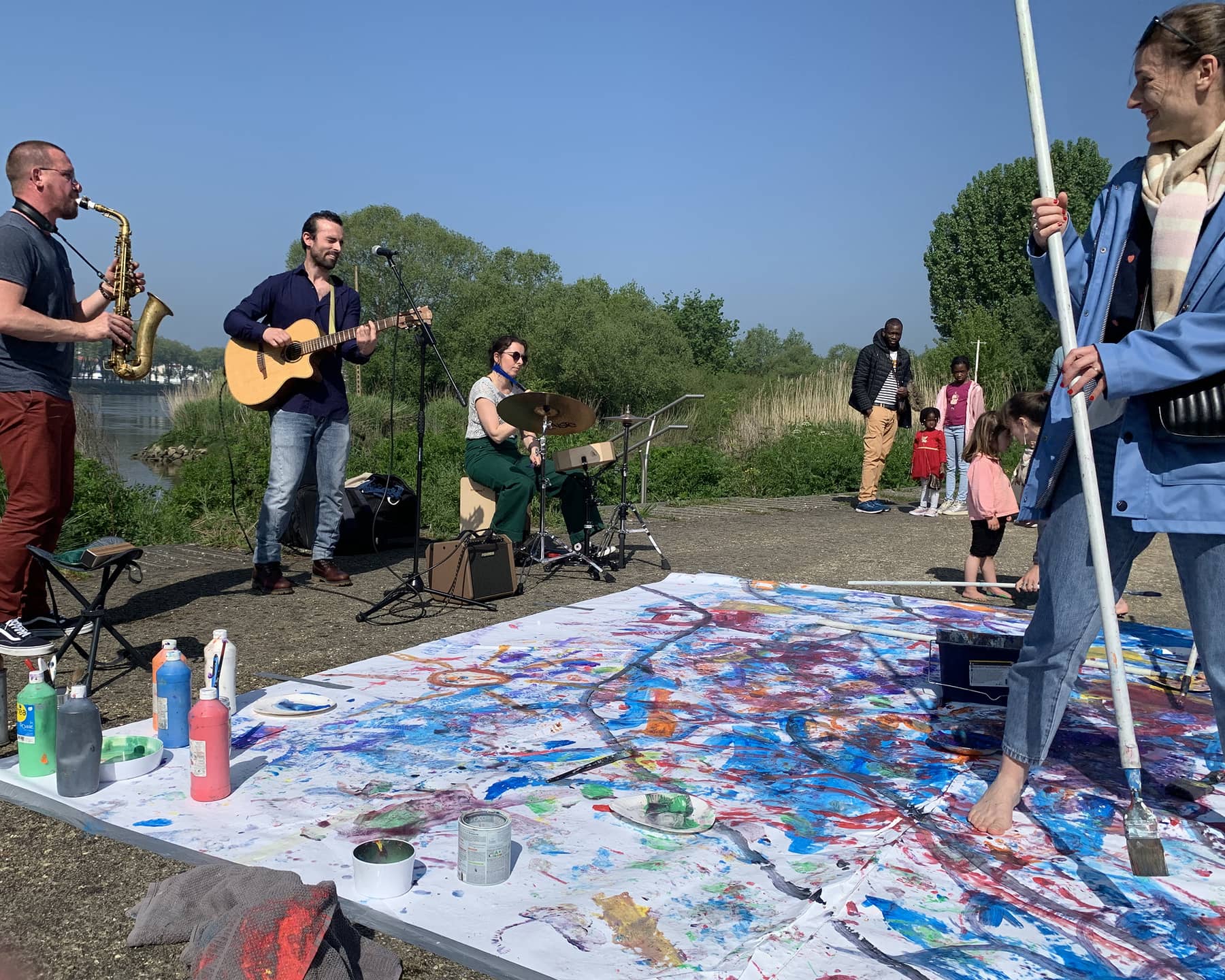 Fresque en papier au sol avec de la peinture. Autour, des spaectateurs et des artistes qui jouent de la musique